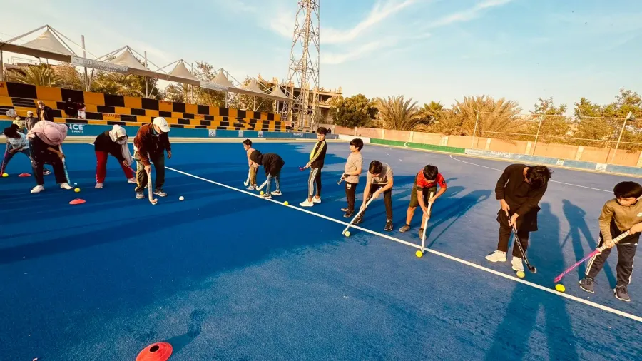 Children and coaches spread in a line across the blue turf, each with a stick and ball